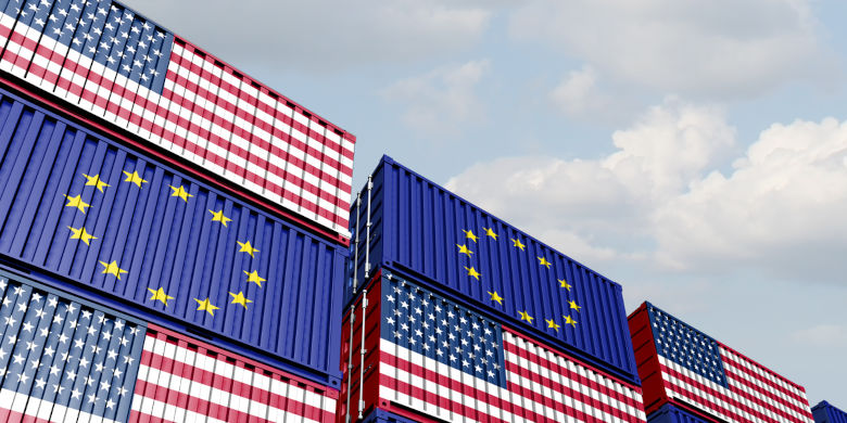 Flera staplade fraktcontainrar målade med EU:s och USA:s flaggor. De står mot en ljus himmel och illustrerar internationell handel mellan EU och USA. Foto.