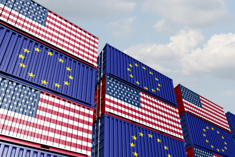 Flera staplade fraktcontainrar målade med EU:s och USA:s flaggor. De står mot en ljus himmel och illustrerar internationell handel mellan EU och USA. Foto.