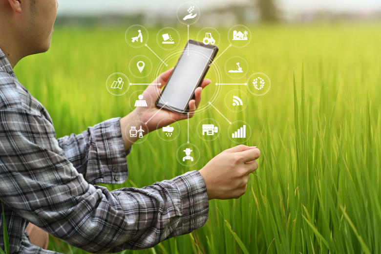En man står framför ett fält och håller en smartphone framför sig, där flera applikationer illustreras bildligt med hjälp av symboler och pilar. Foto.